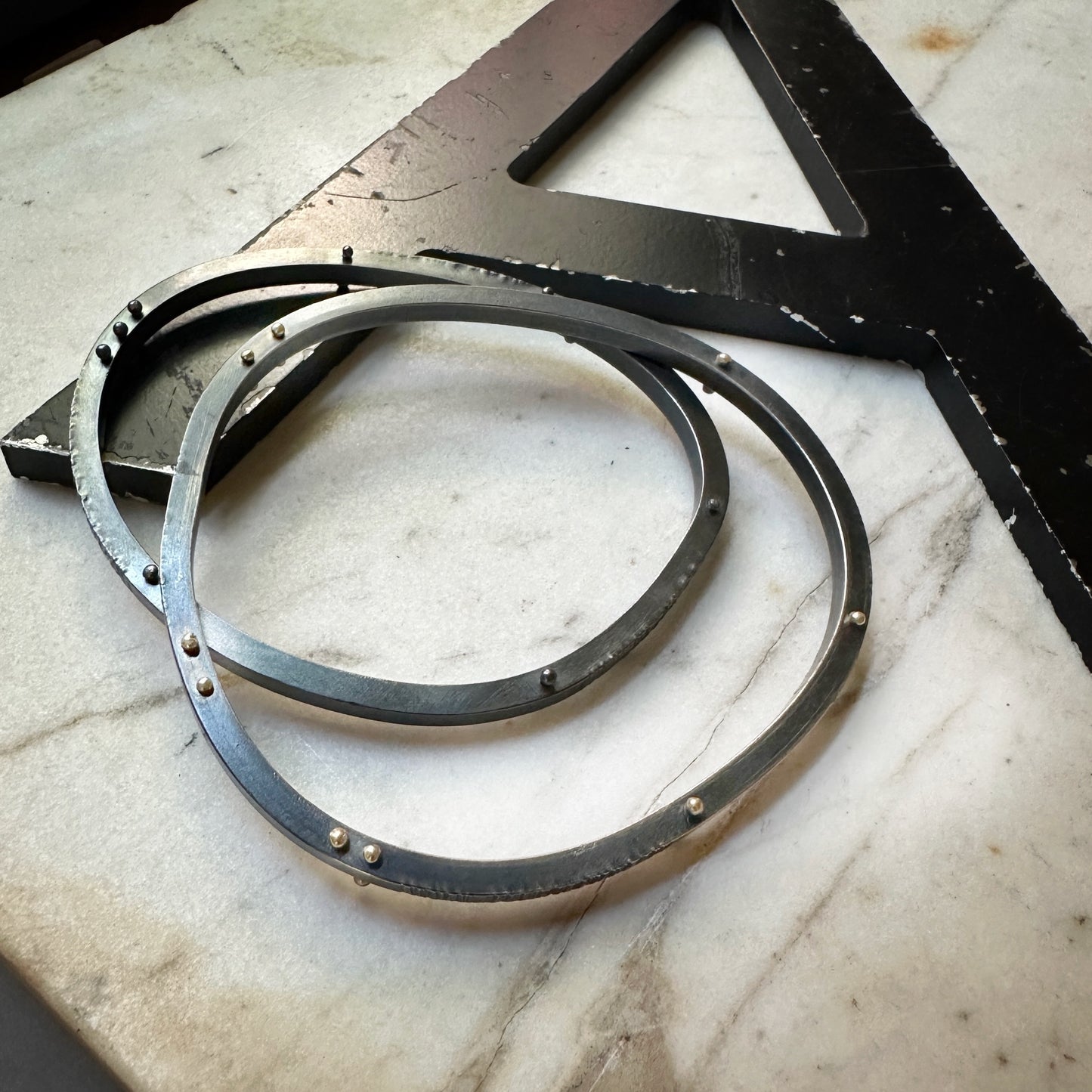 Two oxidized silver bangle bracelets with small rivets, placed on a light marble-like surface next to a large black metal square.