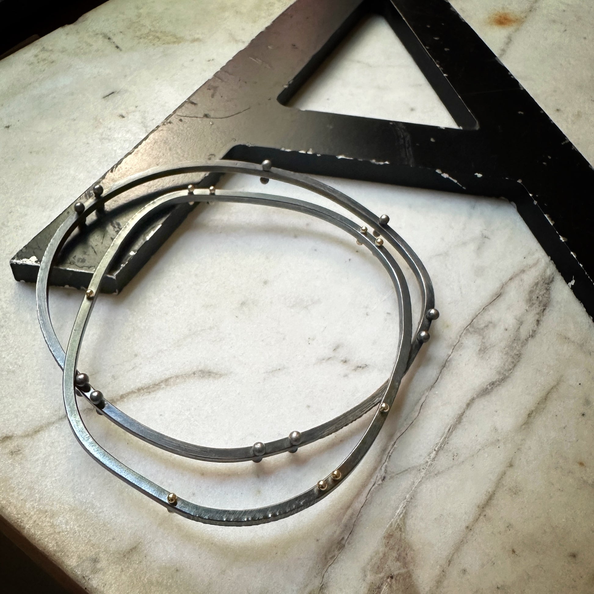 Three thin oxidized silver bangle bracelets with small rivet accents, arranged on a white marble surface next to a black metal square.