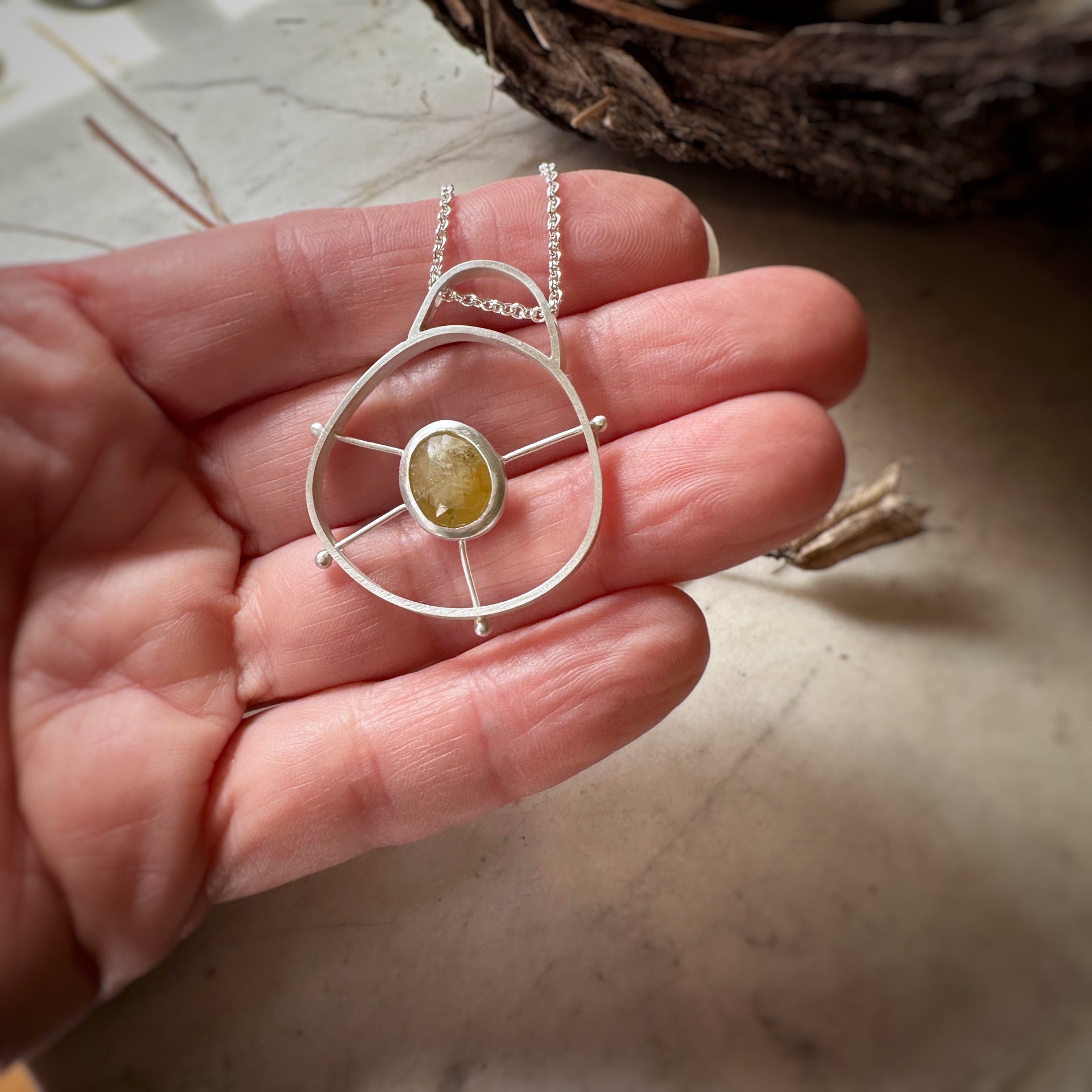 A sterling silver circular pendant with a domed top. There is a bezel set yellow sapphire suspended in the middle of the circle by 4 posts.  Shown on a hand for scale