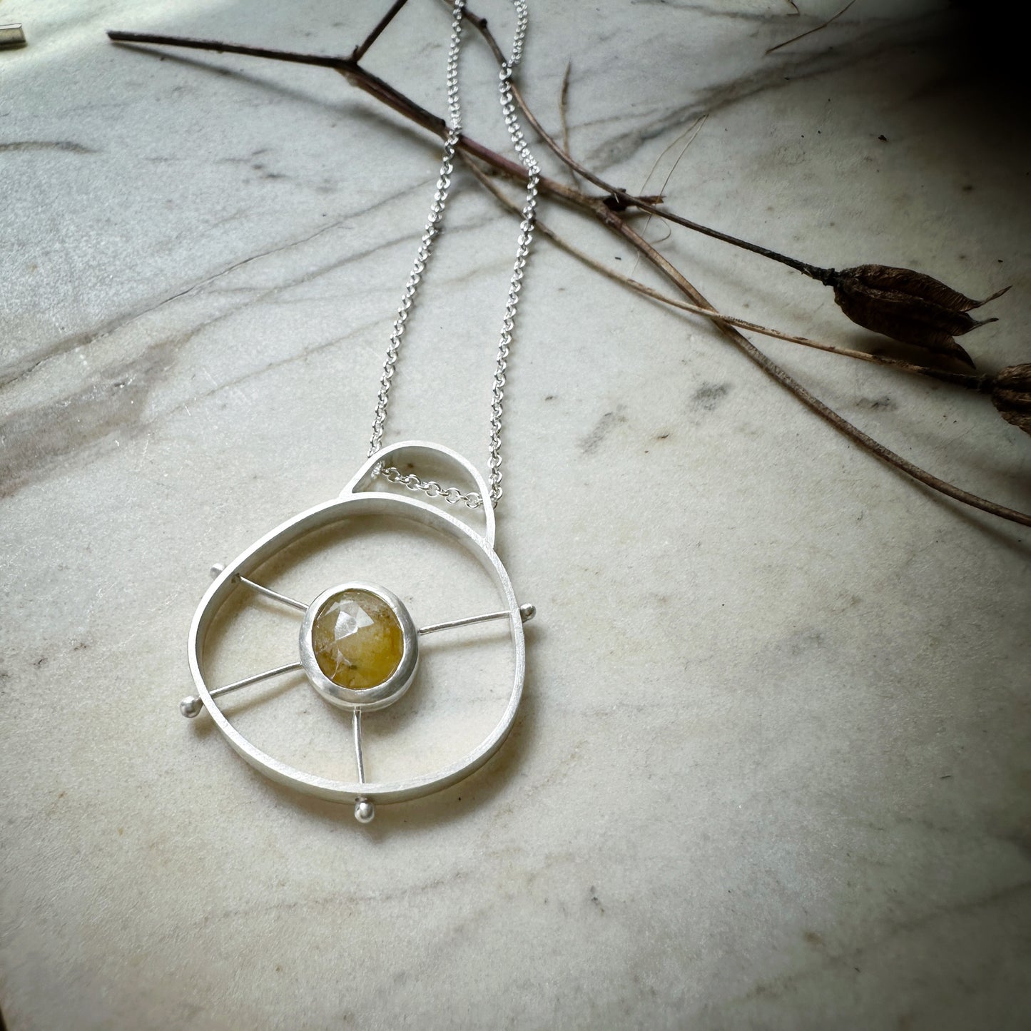 A sterling silver circular pendant with a domed top.  There is a bezel set yellow sapphire suspended in the middle of the circle by 4 posts.