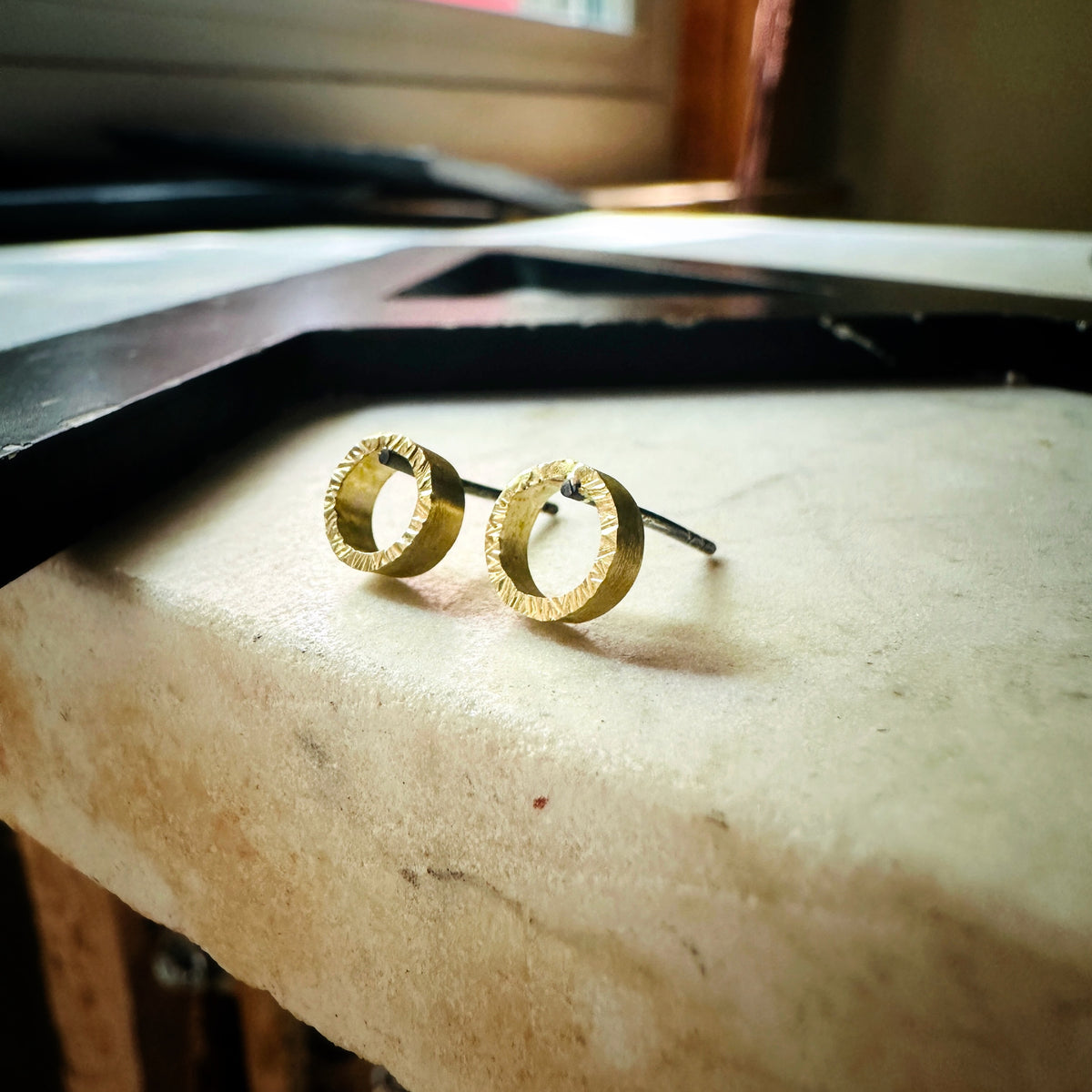 A pair of textured gold hoop earrings resting on a light beige stone surface, with a dark tray and a softly blurred indoor background.