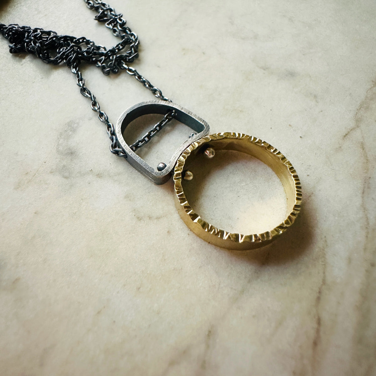a hammered gold circle pendant with a oxidized silver chain and bail on a marble surface
