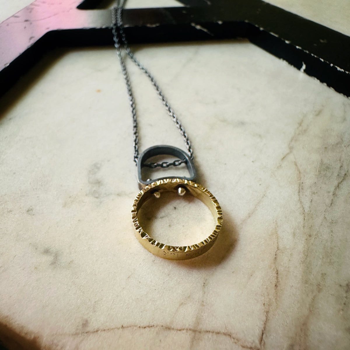 A gold textured ring pendant necklace with a silver metal connector and chain, resting on a beige marble surface.