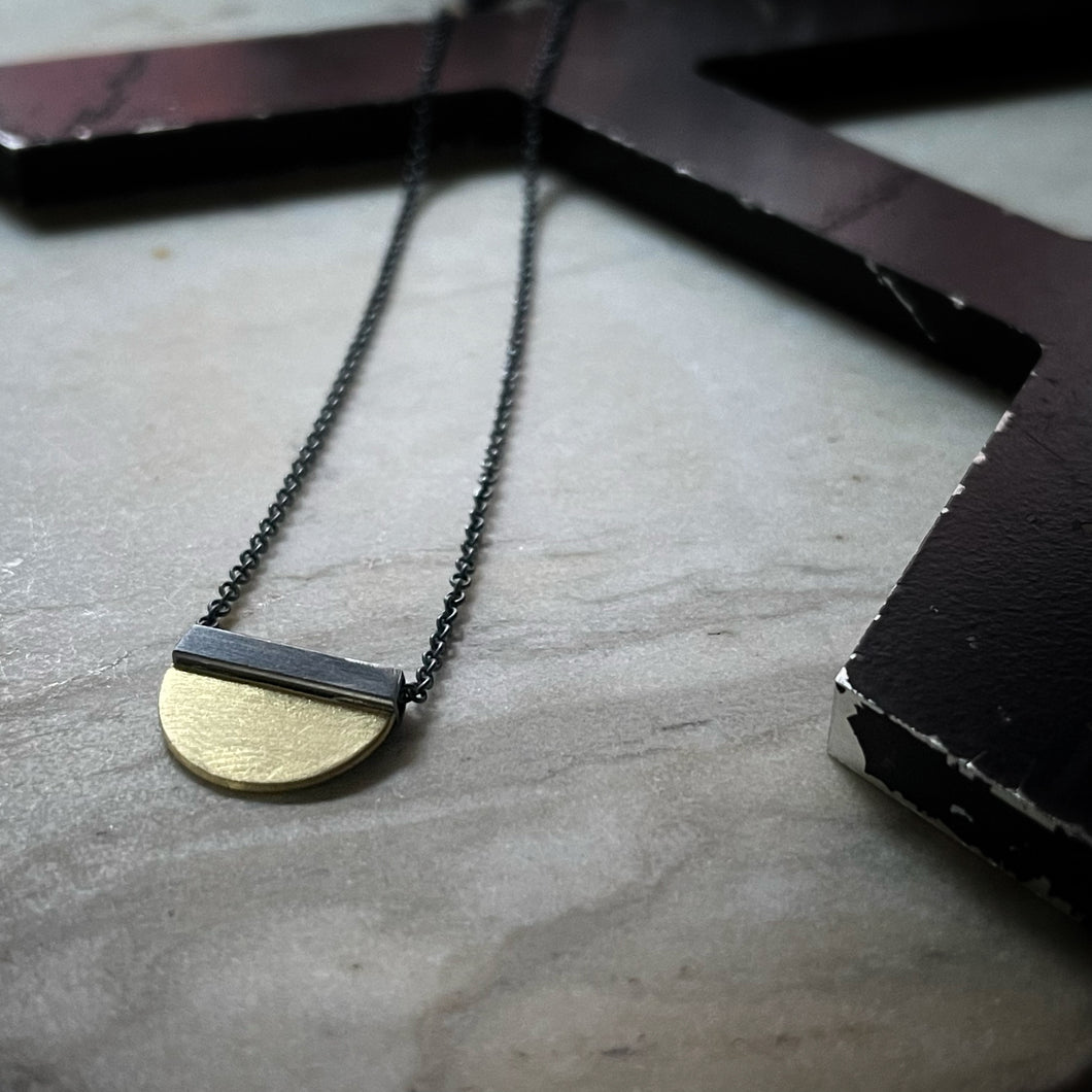 a half circle of yellow gold soldered to a oxidized silver square bail.  An oxidized chain runs through the bail. 