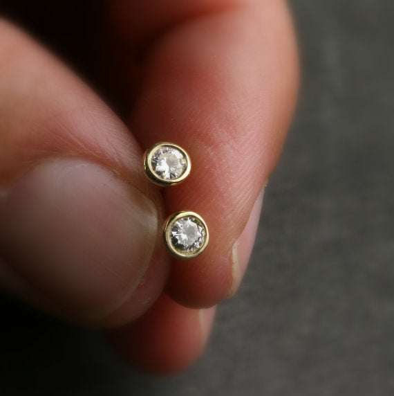 A hand holding two 18k yellow gold bezel set earrings with 3mm Moissanite stones