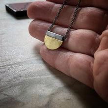 Load image into Gallery viewer, a half circle of yellow gold soldered to a oxidized silver square bail.  An oxidized chain runs through the bail.  Shown on a hand for scale
