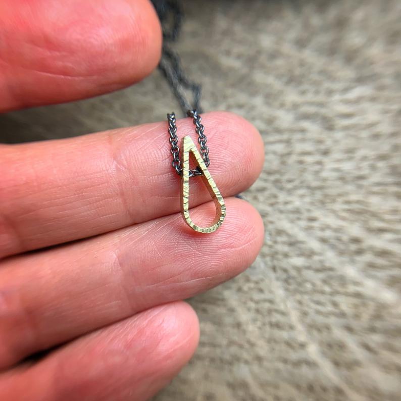 tiny hammered teardrop shaped pendant in 18k yellow gold on an oxidized sterling silver chain.  Shown against a hand for scale.