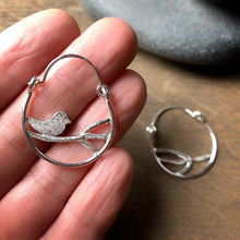 Load image into Gallery viewer, two hoop earrings in sterling silver.  Both have textured branches inside the hoop and there is one bird sitting on the branch.  One earring is on a hand for scale
