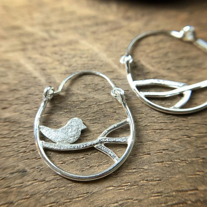 two hoop earrings in sterling silver.  Both have textured branches inside the hoop and there is one bird sitting on the branch. 