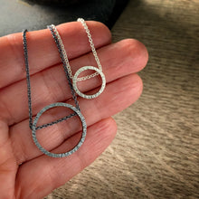 Load image into Gallery viewer, two pendants, one large and one small, in sterling silver.  They are circles with a hammered texture and the chain runs through the circle.  One is darkened silver and the other bright silver.  Shown on a hand for scale
