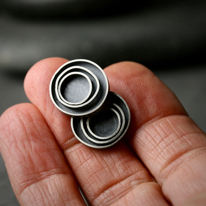 Oxidized sterling silver concentric circle post earrings shown on a hand for scale